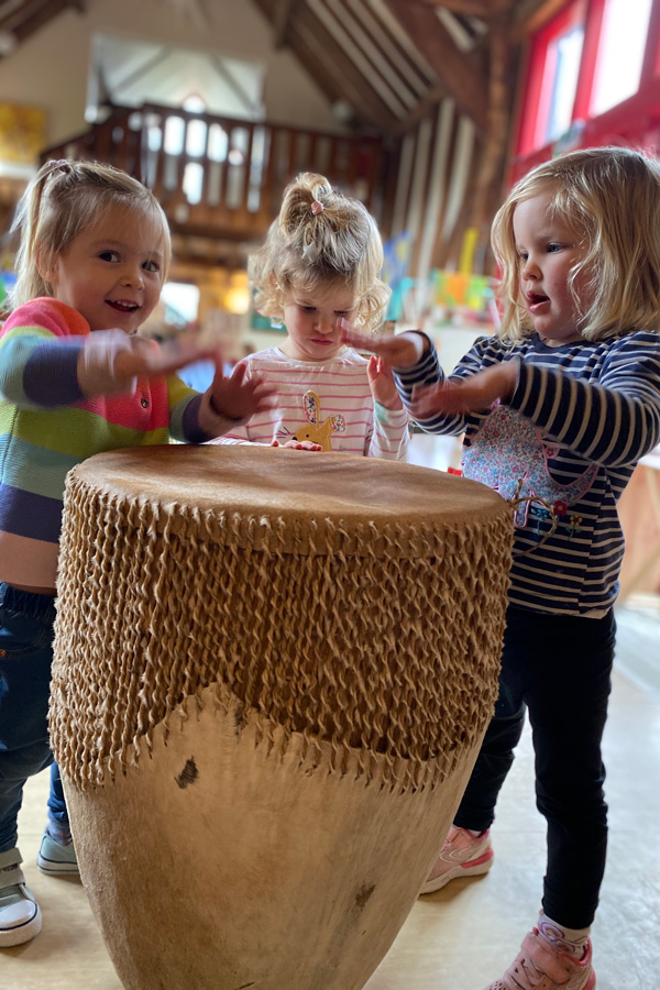 Three young children surrounding and patting a large drum (Grantham Farm Montessori School)